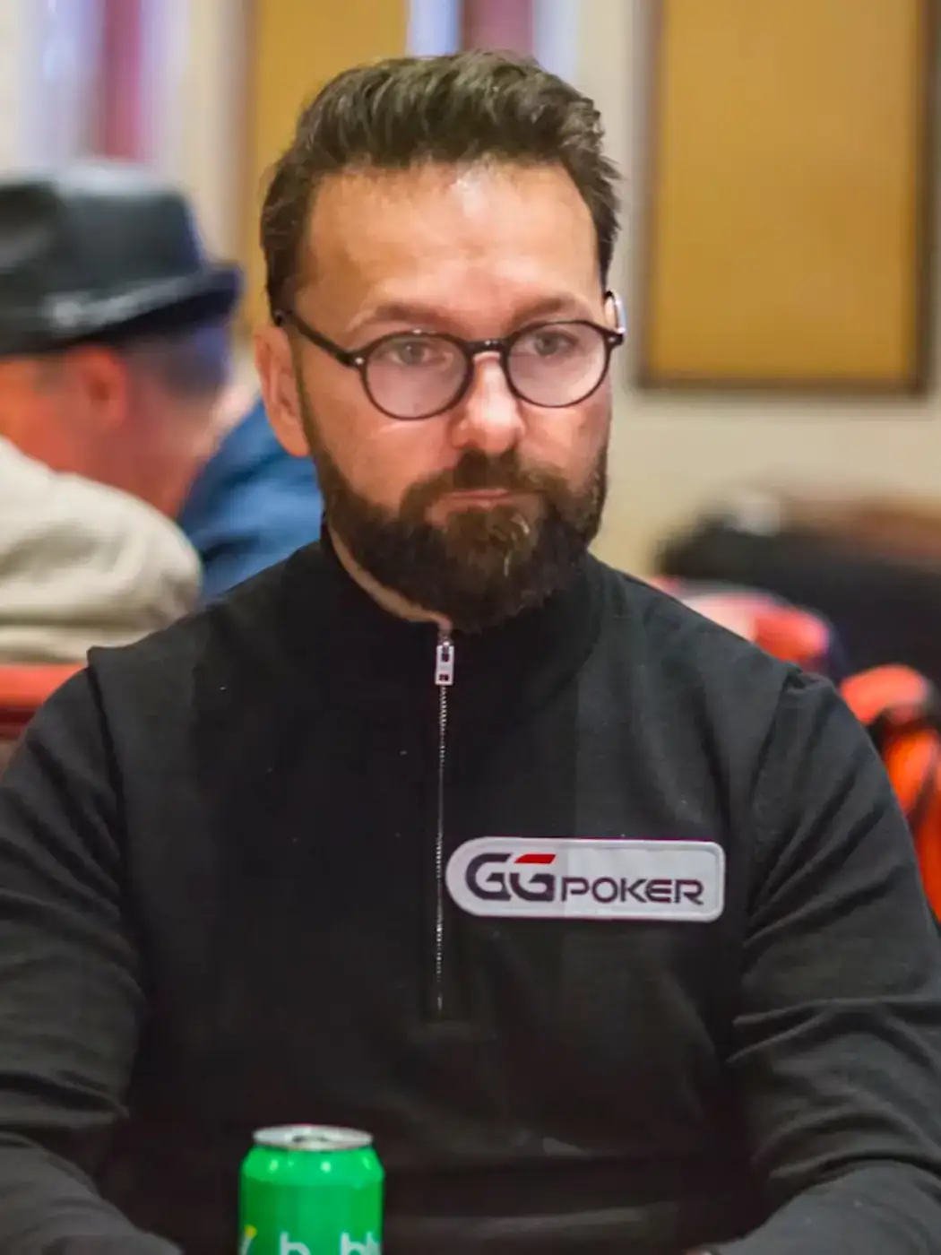Daniel Negreanu seated at a poker table in 2019 wearing glasses and a dark zip top, his hair combed back off the forehead under room lighting.