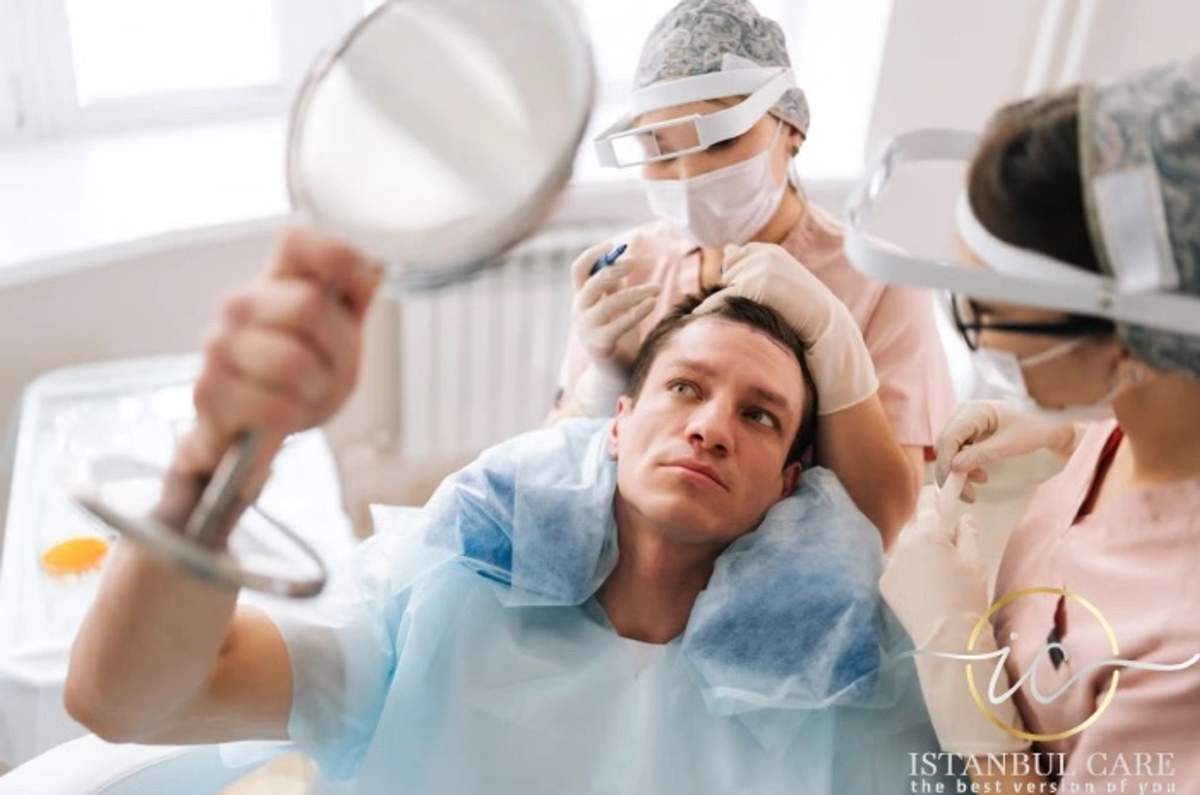 An unidentified patient in a surgical cap holding a hand mirror up to his own face while two clinicians work around him.