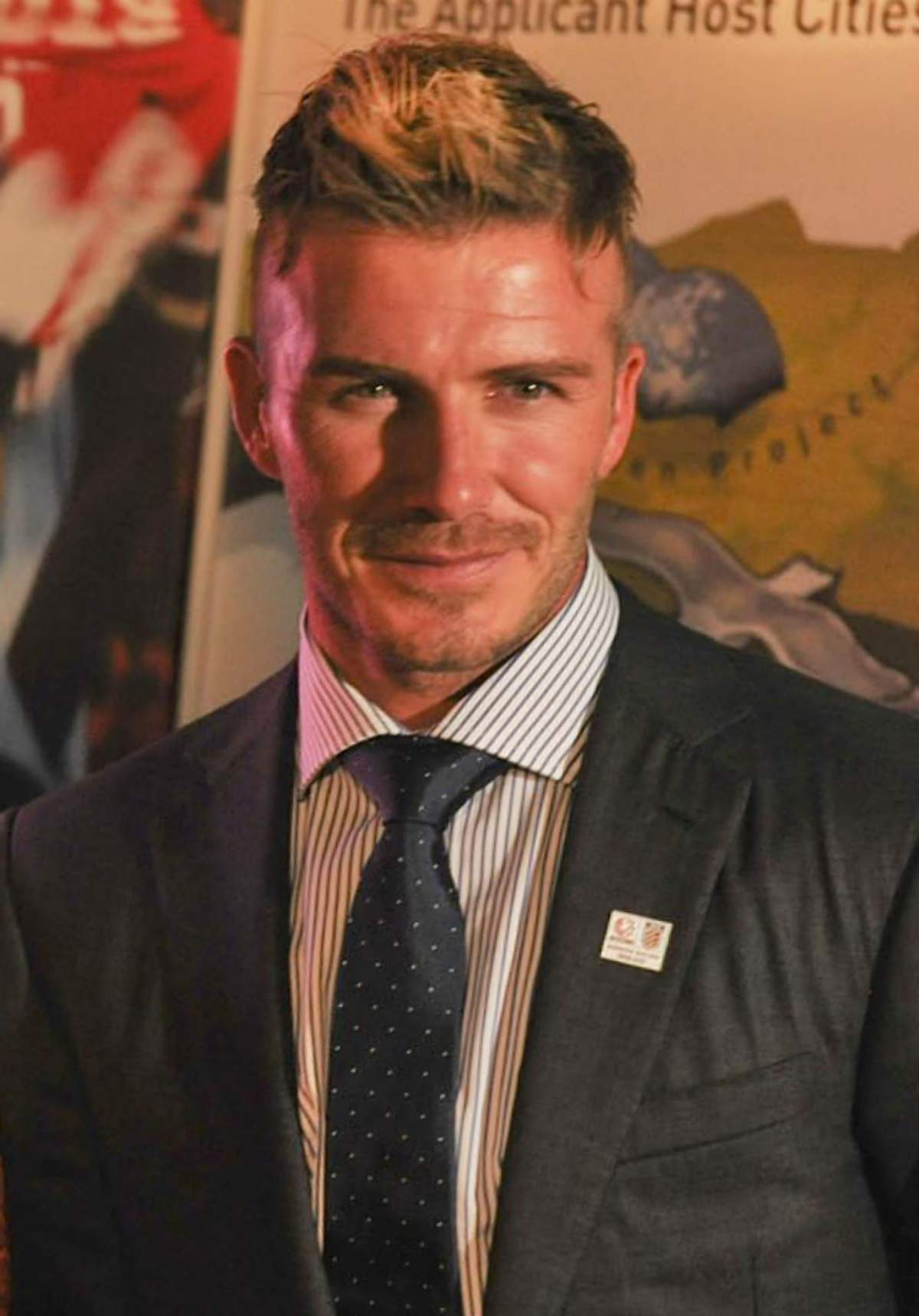 David Beckham in a suit at an indoor press event, photographed front-on with his hair cut short at the sides and styled upward at the front.
