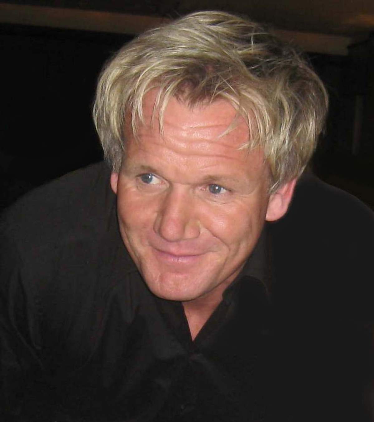 Gordon Ramsay at an indoor event photographed front-on under hard flash, his light blond hair pushed up and back from the forehead.