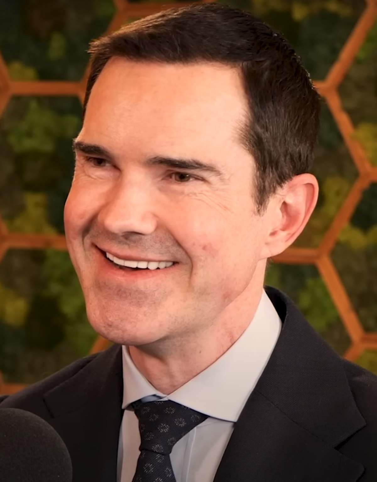 Jimmy Carr sonriendo con traje en un evento al aire libre en 2024, su cabello oscuro peinado hacia atrás y la línea frontal del cabello visible a la luz del día.