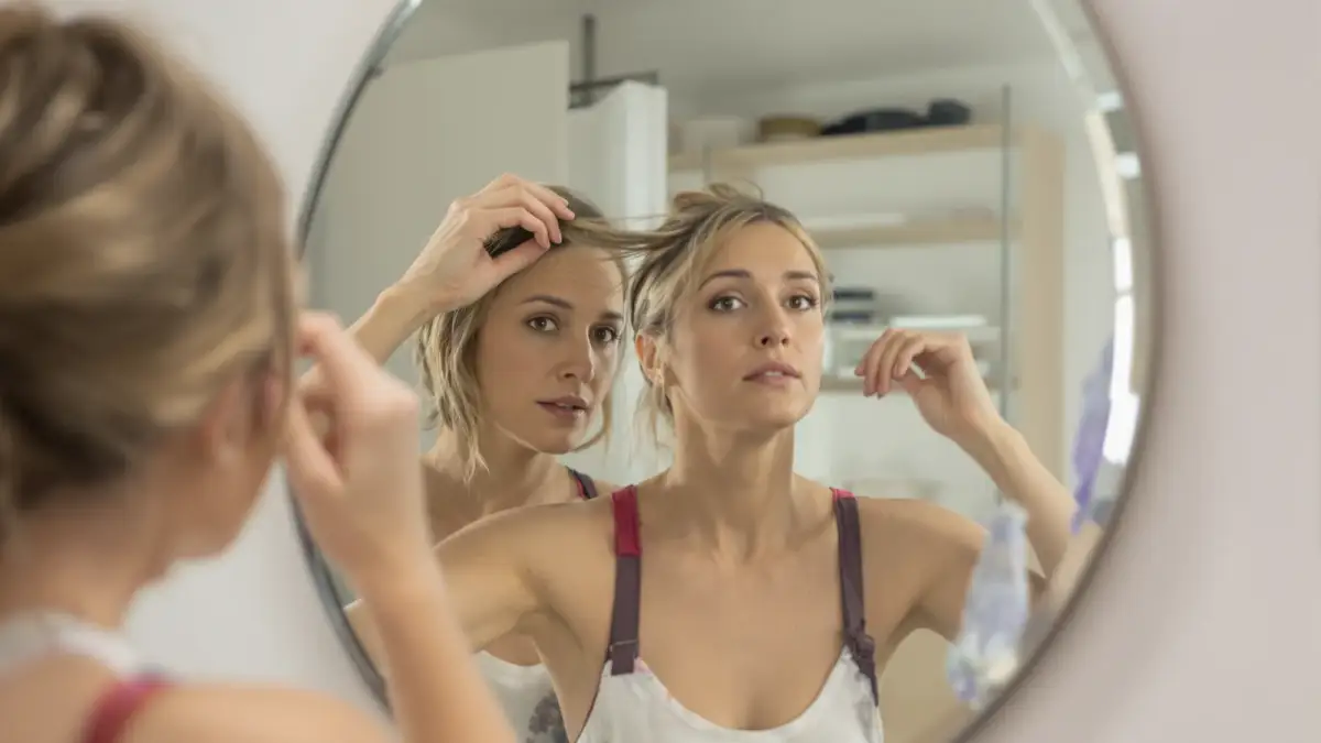Woman checking her hair parting in a bathroom mirror while assessing thinning at the crown, illustrating how women evaluate hair regrowth products such as Keranique