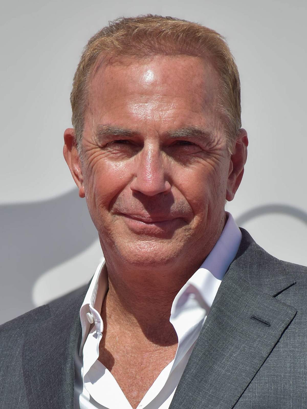 Kevin Costner en un festival de cine de 2024 con chaqueta gris, fotografiado de frente con luz plana de prensa y el cabello peinado hacia un lado sobre la frente.
