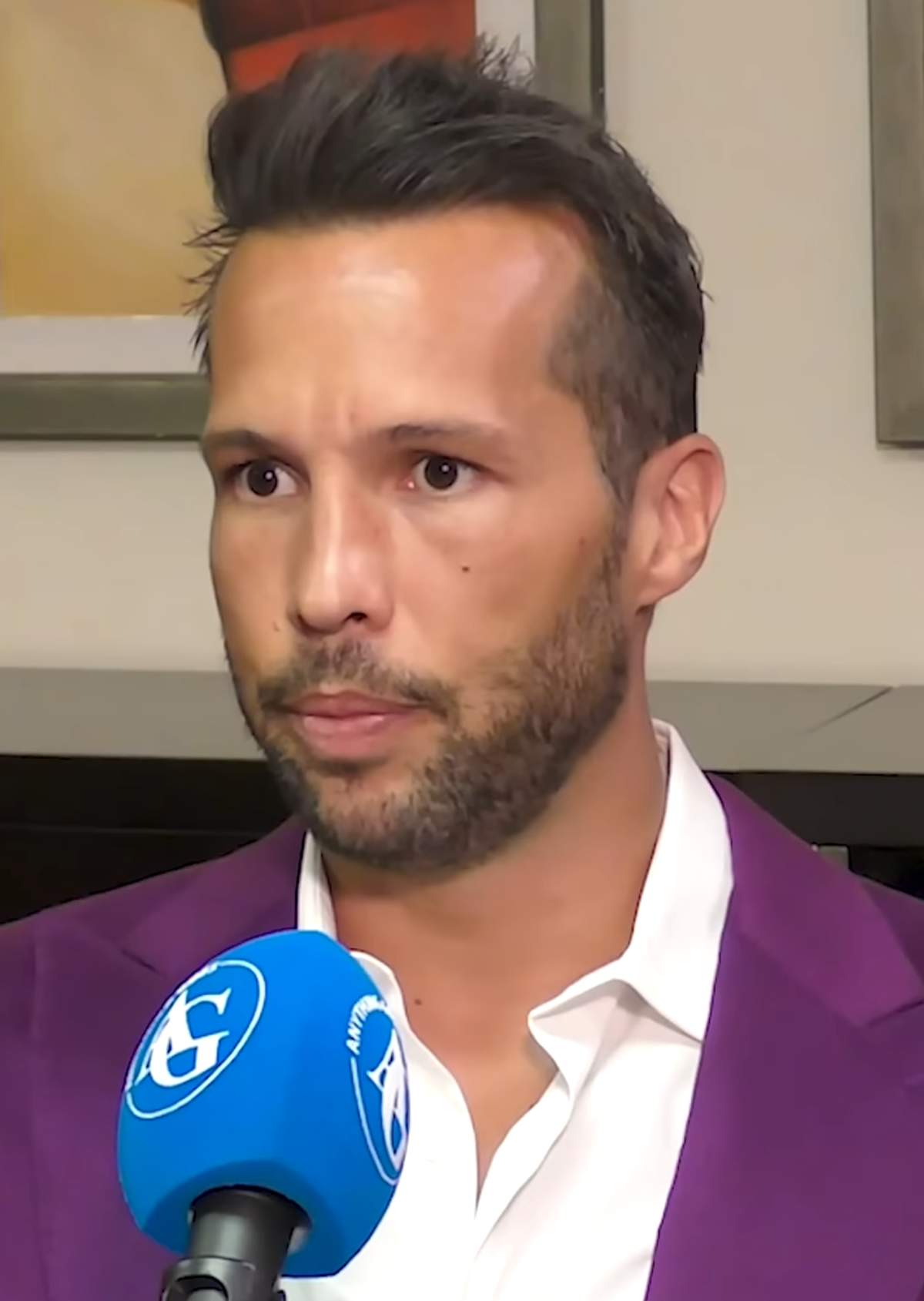 Tristan Tate speaking into a branded interview microphone indoors, his hair styled up and back from the forehead under studio light.