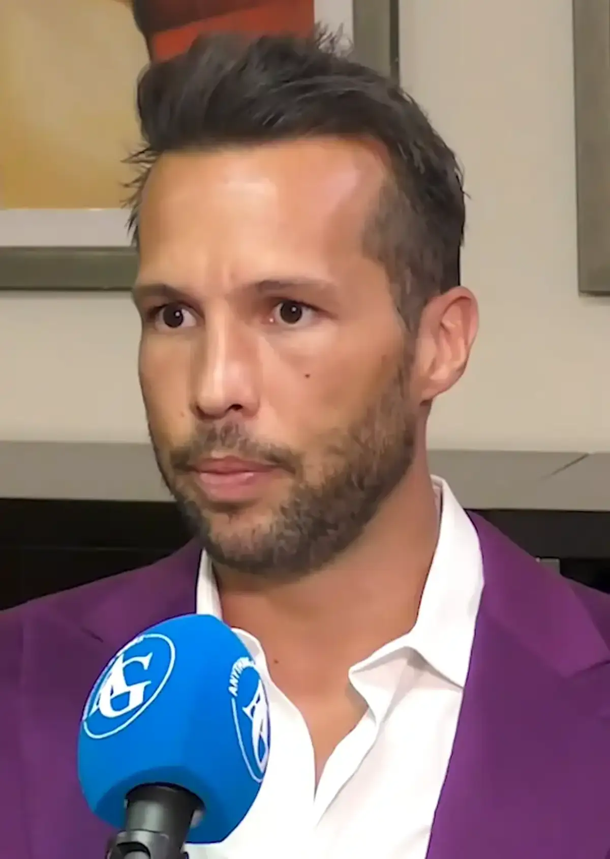 Tristan Tate speaking into a branded interview microphone indoors, his hair styled up and back from the forehead under studio light.