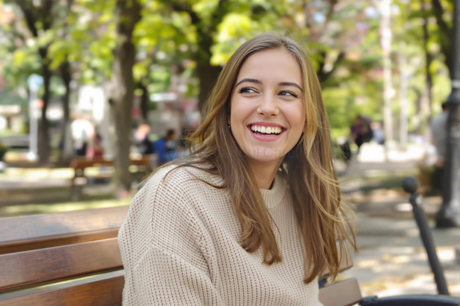 Woman with long fair hair sitting on a park bench in daylight, laughing with her upper front teeth visible.