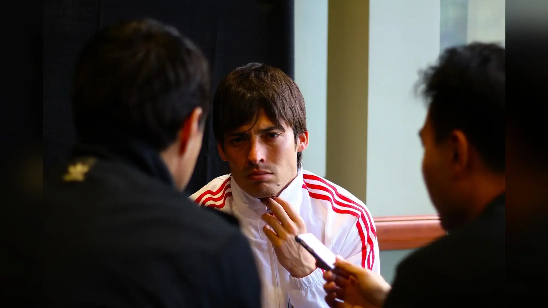 David Silva in a white and red Spain tracksuit at a press table, photographed head-on with his dark hair and front hairline in view.