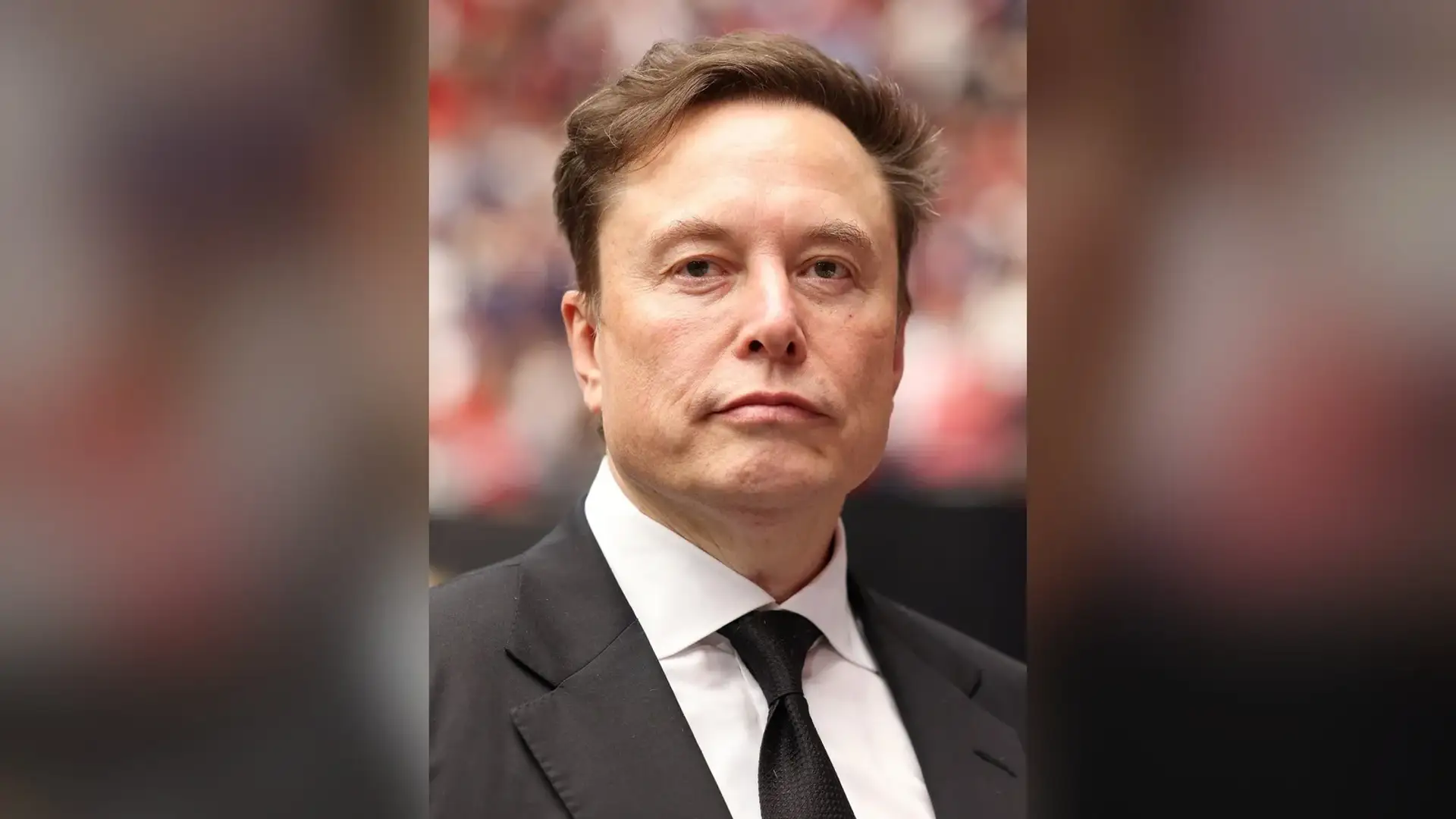 Elon Musk in a dark suit and tie photographed from the front at a public event, with his forehead and front hairline in view.