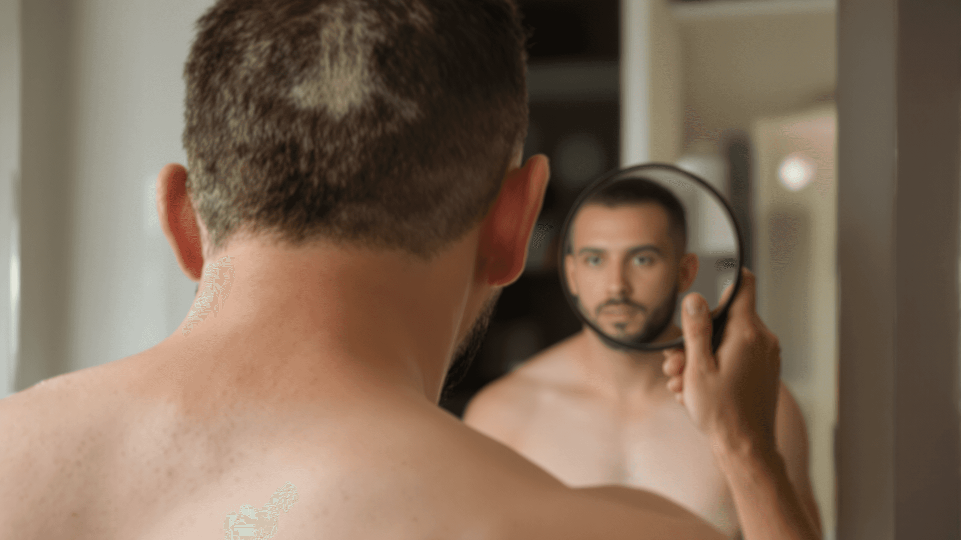 Man examining the back of his scalp with a hand mirror, donor area visible after a hair transplant.