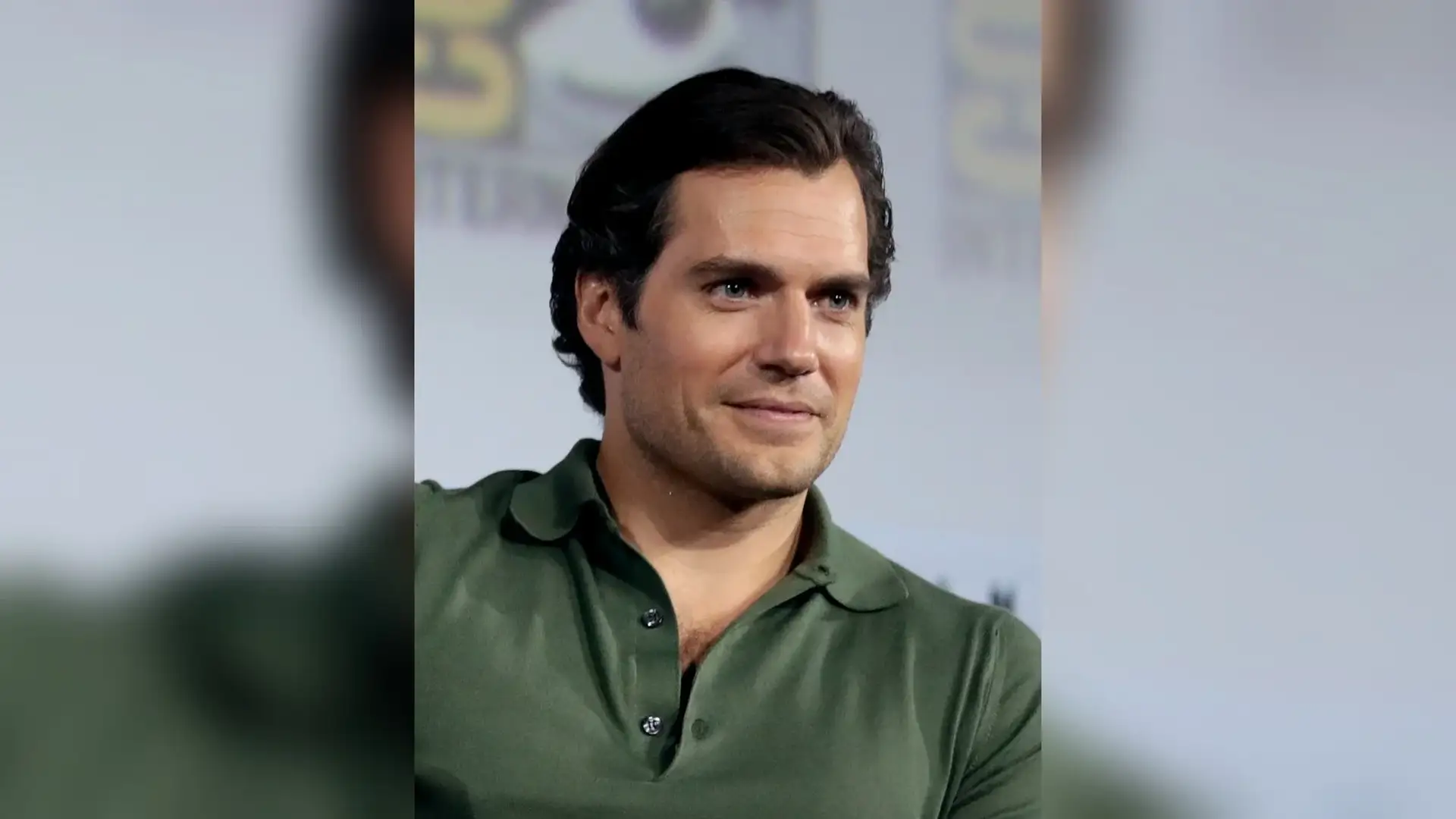 Henry Cavill in a green polo shirt at a convention panel, head turned slightly aside with dark hair swept back off his forehead.