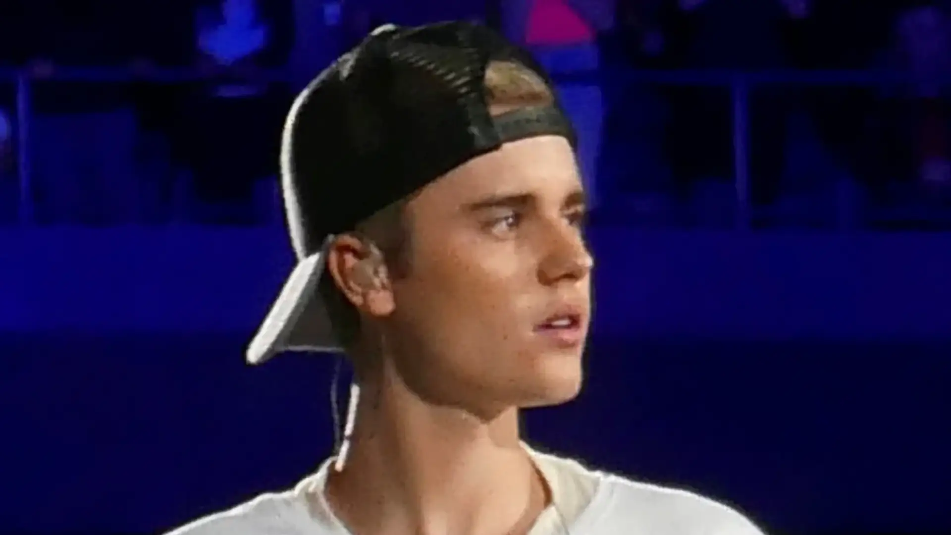 Justin Bieber performing on stage in a white T-shirt and a backwards cap, seen in three-quarter profile with his temple and front hairline visible.