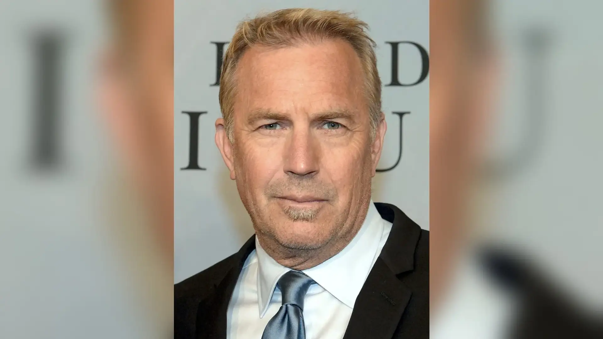 Kevin Costner in a dark suit and pale blue tie at a press event, facing the camera with his forehead and full hairline in view.