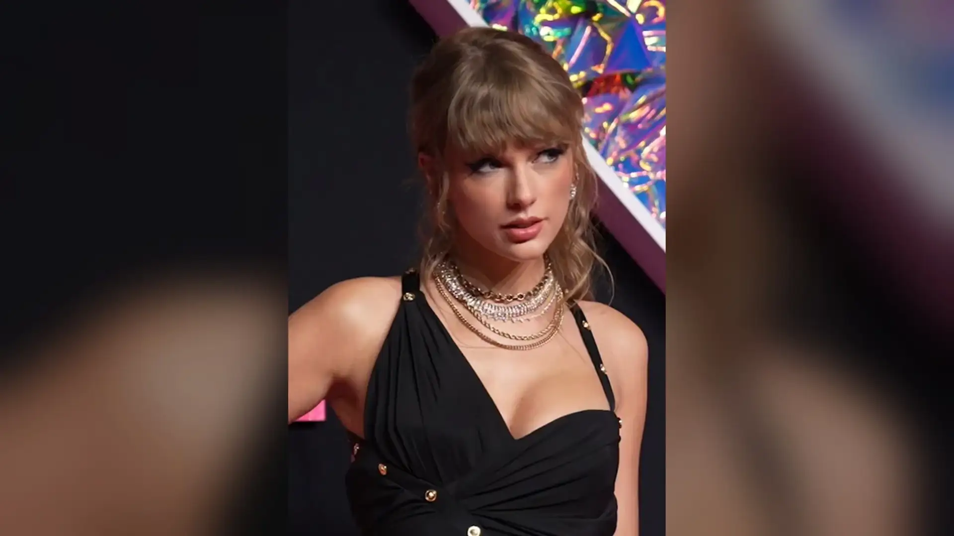 Taylor Swift on a red carpet in a black dress and layered necklaces, photographed close in three-quarter view.