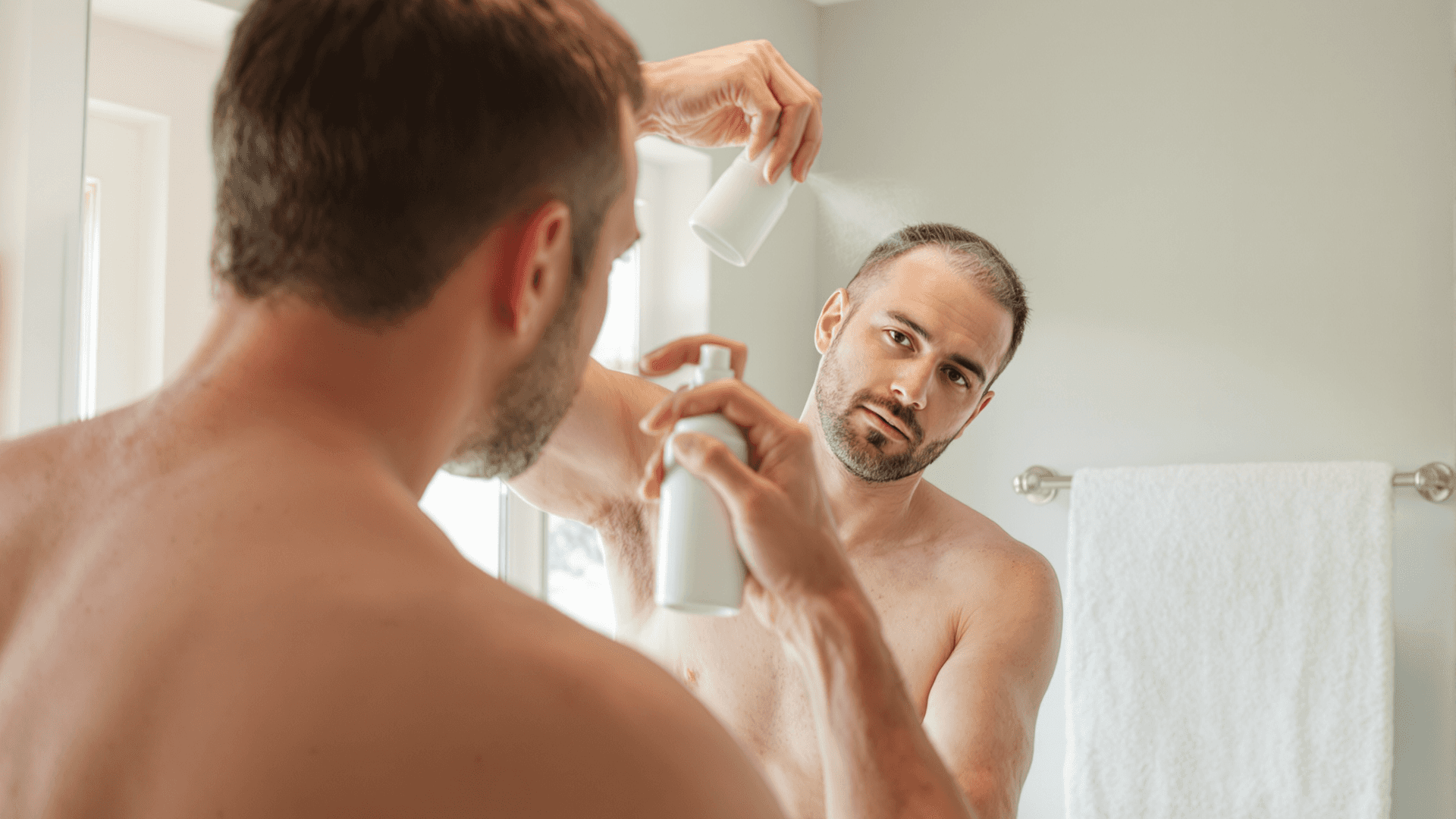 Man applying a spray to his scalp in front of a bathroom mirror after a hair transplant.