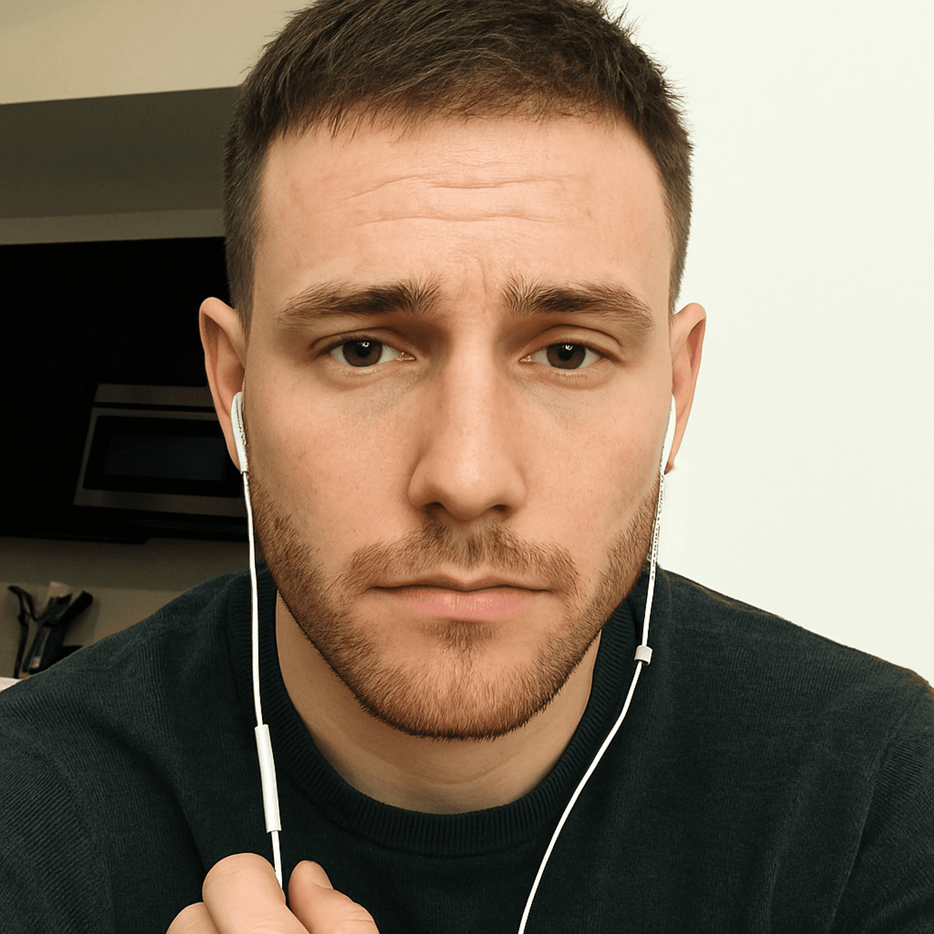 Young man with earbuds, serious expression, wearing a dark sweater, sitting indoors.