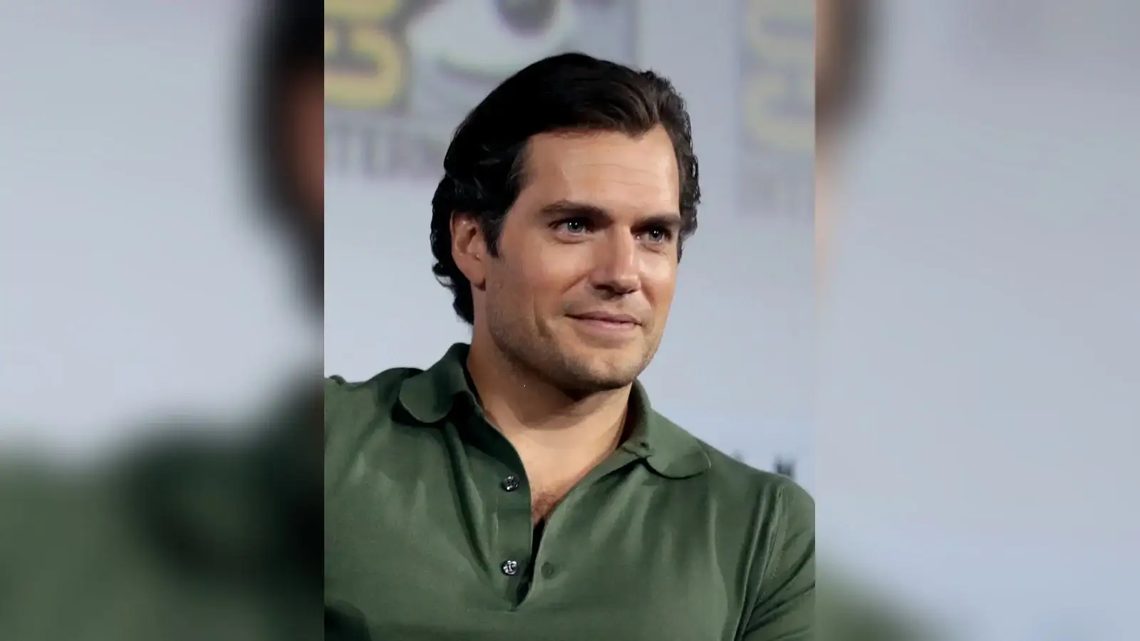 Henry Cavill in a green polo shirt at a convention panel, head turned slightly aside with dark hair swept back off his forehead.