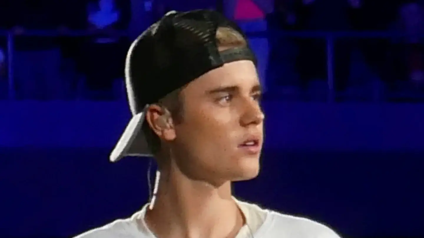 Justin Bieber performing on stage in a white T-shirt and a backwards cap, seen in three-quarter profile with his temple and front hairline visible.