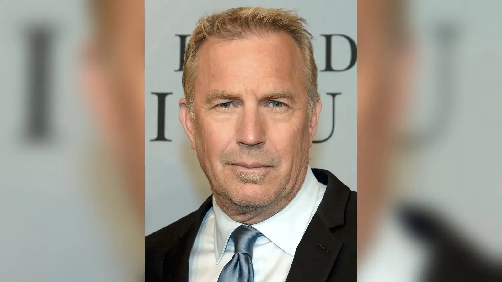 Kevin Costner in a dark suit and pale blue tie at a press event, facing the camera with his forehead and full hairline in view.