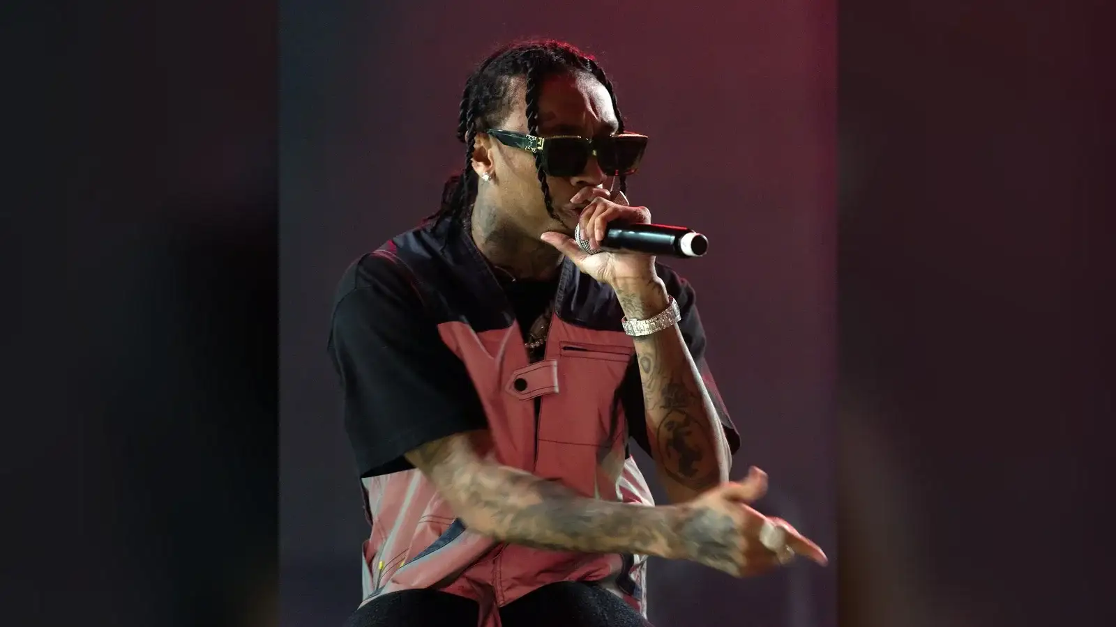 Tyga performing on stage in sunglasses, holding a microphone, with braided hair pulled back from the forehead.