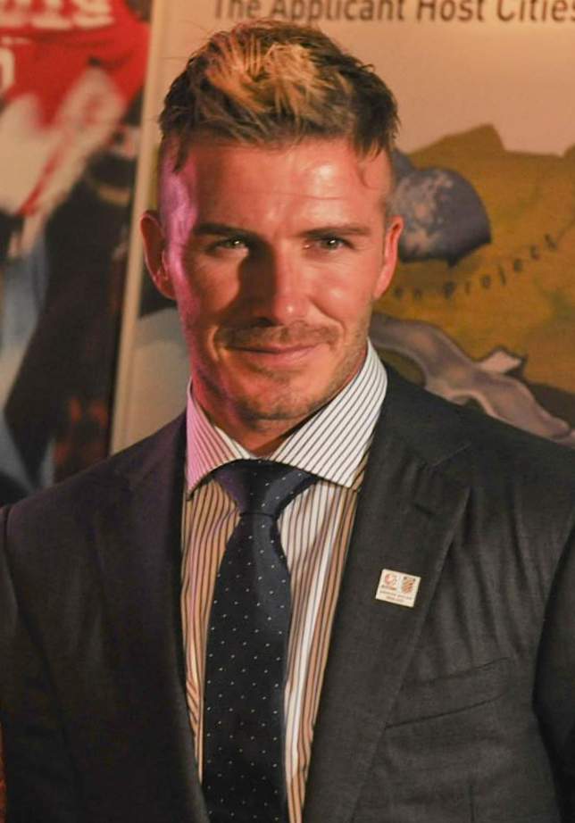 David Beckham in a suit at an indoor press event, photographed front-on with his hair cut short at the sides and styled upward at the front.