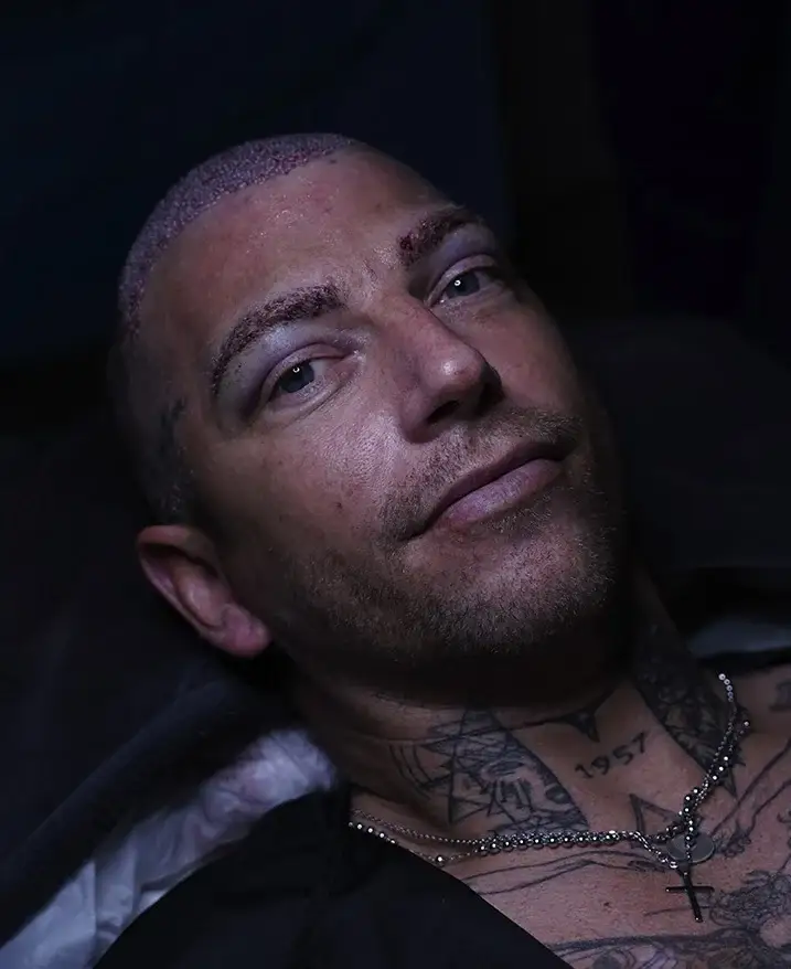 An unidentified man with a closely shaved head and neck tattoos, photographed reclining against a dark background.