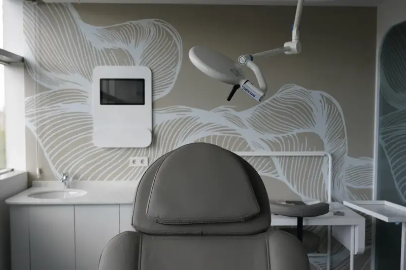 View over a treatment chair headrest towards a surgical light, a wall-mounted screen and a washbasin
