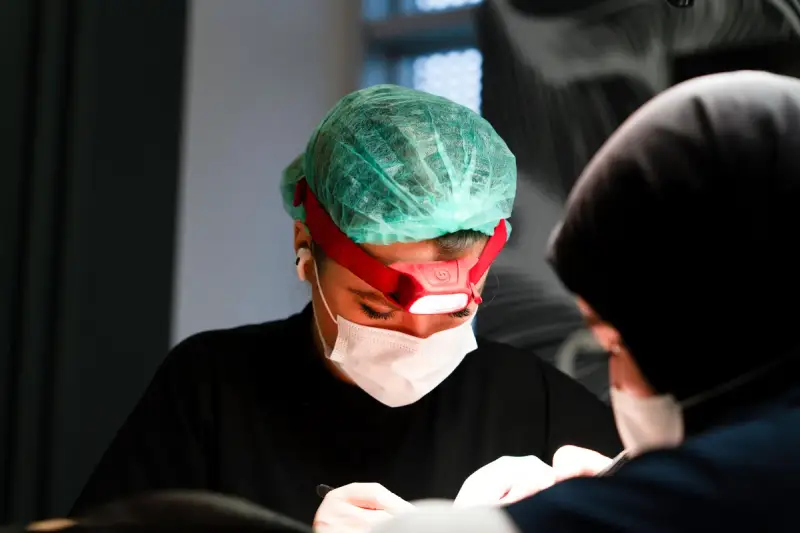 Two staff members in surgical masks at work during a procedure, one wearing a cap and a headlamp