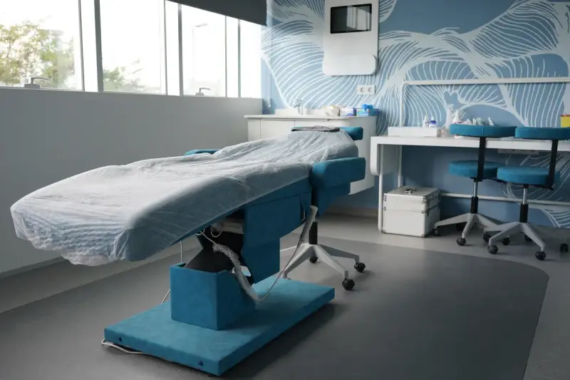 Treatment bed with a disposable cover in a daylit room, next to a work table and two stools