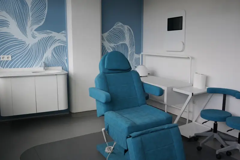 Treatment room with a blue reclining chair, a washbasin, a wall-mounted screen and stools