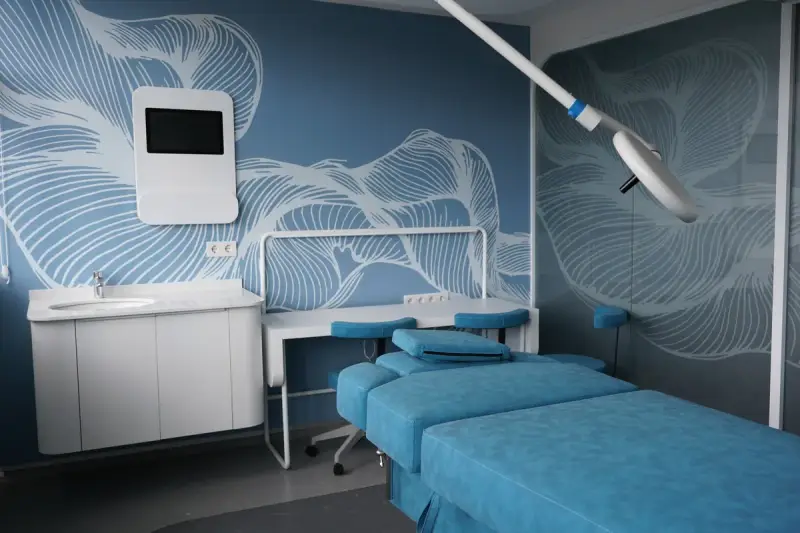 Blue treatment bed under an examination light on a ceiling arm, with a washbasin and a wall screen