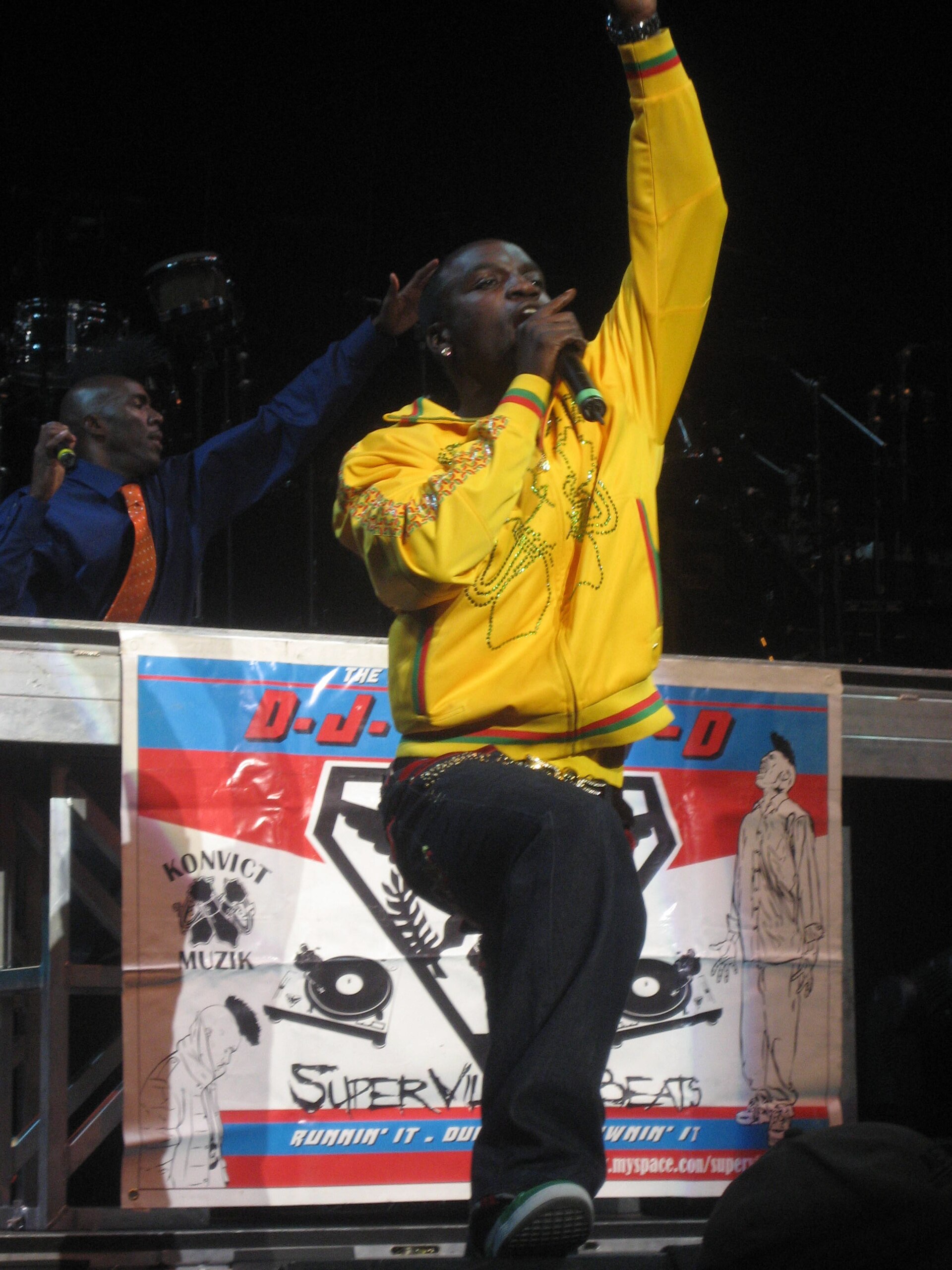 Akon si esibisce sul palco in una tuta gialla, fotografato dal pubblico sotto il livello del palco, con la testa piccola nell'inquadratura e l'attaccatura dei capelli non leggibile.