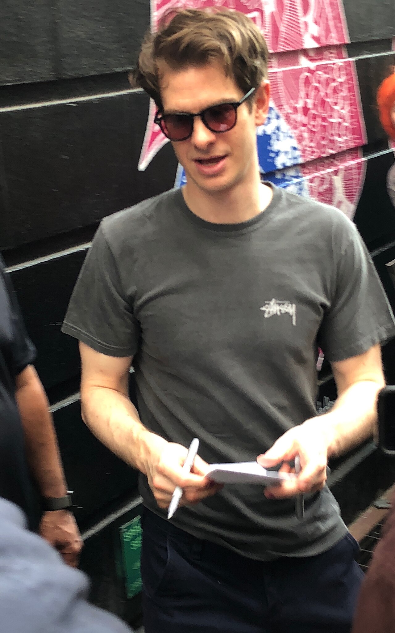 Andrew Garfield outdoors in sunglasses and a grey T-shirt, his hair worn short and pushed back so the temple corners are visible.