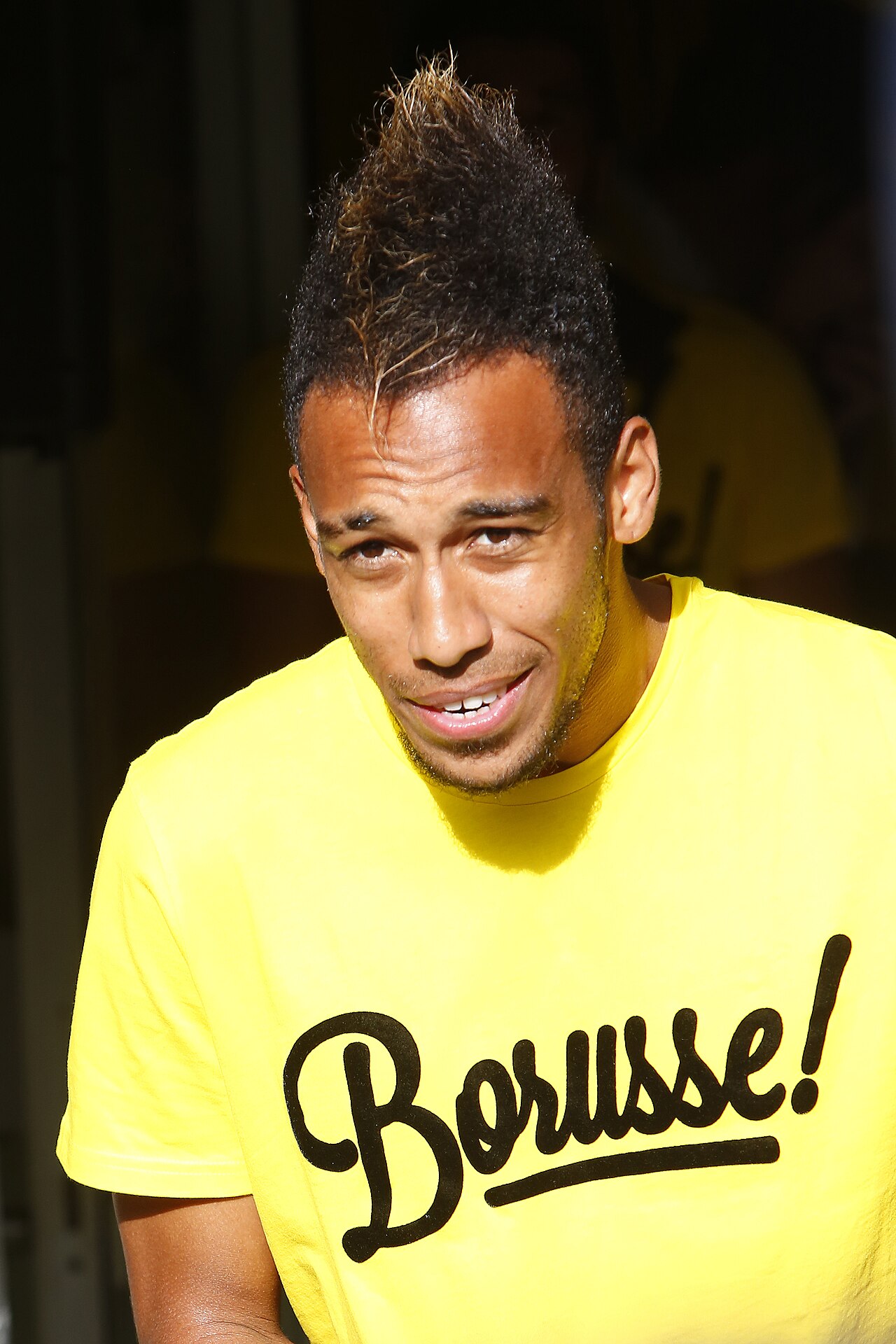 Pierre-Emerick Aubameyang in una maglia gialla del Borussia Dortmund nel 2014, con i capelli pettinati all'indietro in uno stile alto che solleva l'intera sezione frontale dalla fronte.