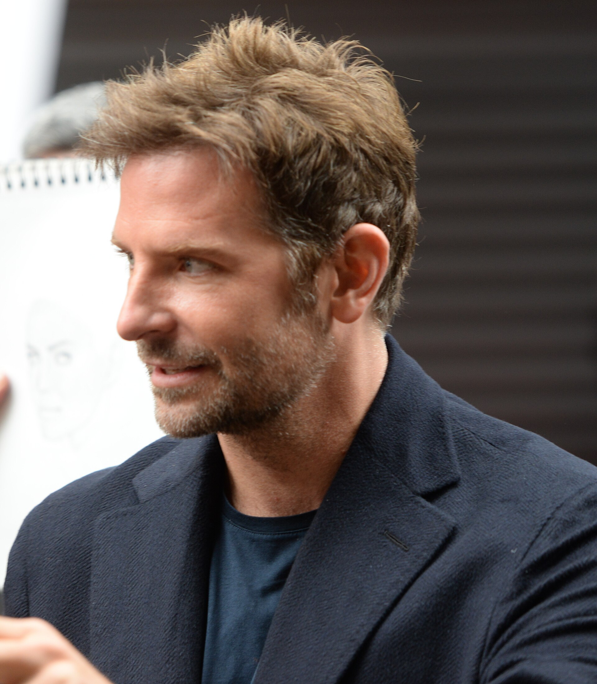 Bradley Cooper fotografato di profilo a un'apparizione pubblica del 2018, con i capelli corti ai lati e lunghi sulla parte superiore, pettinati all'indietro lontano dal viso.
