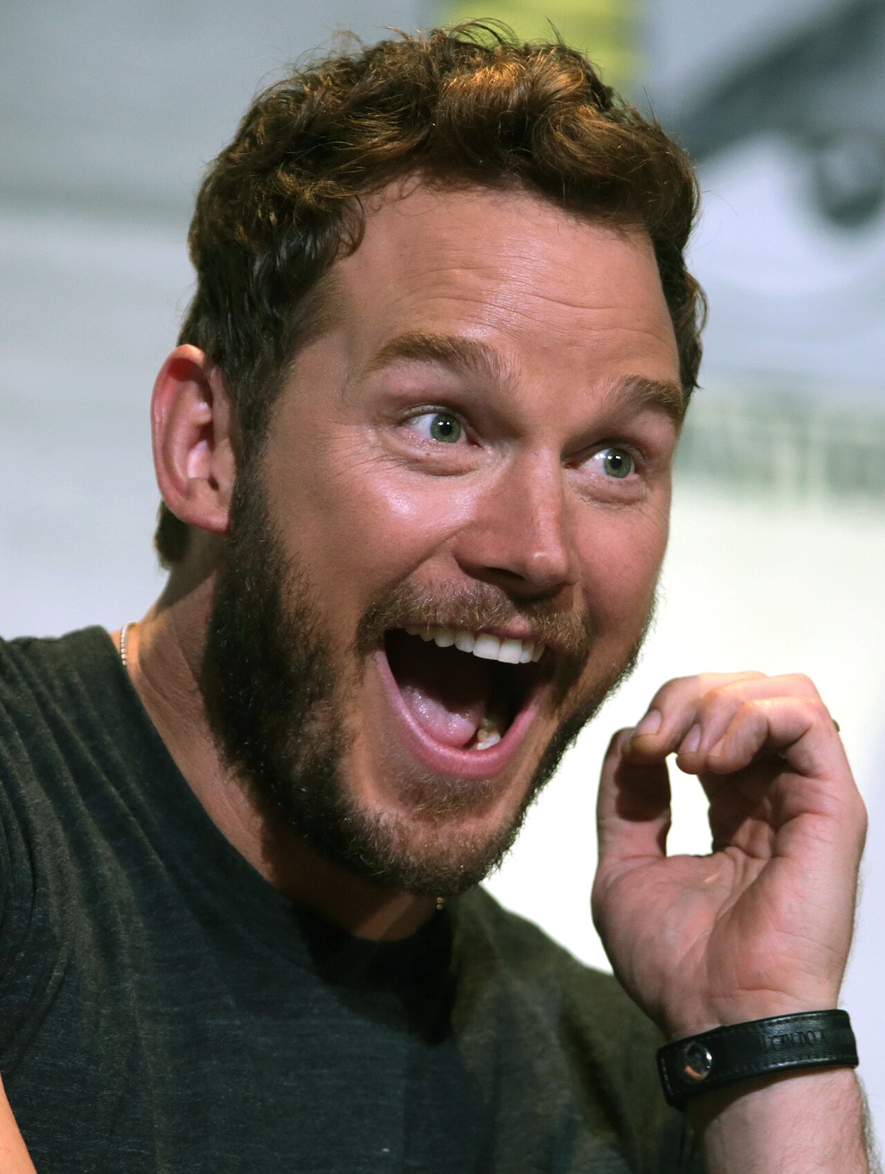 Chris Pratt at a convention laughing with his head turned, his hair swept back off the forehead and the front hairline lit from the side.