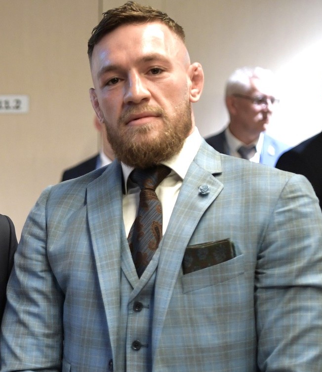 Conor McGregor in un abito a tre pezzi a quadri durante un evento stampa al chiuso nel 2018, con i capelli tagliati corti e le tempie rasate, e l'attaccatura frontale visibile sotto la luce dall'alto.