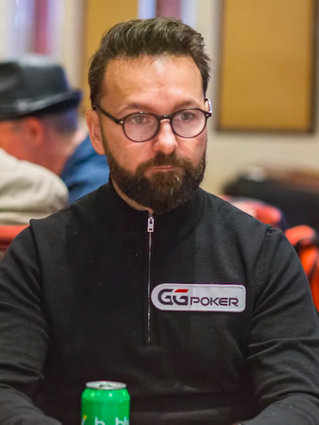 Daniel Negreanu seduto a un tavolo da poker nel 2019 con occhiali e una felpa scura con zip, i capelli pettinati all'indietro dalla fronte sotto la luce della stanza.
