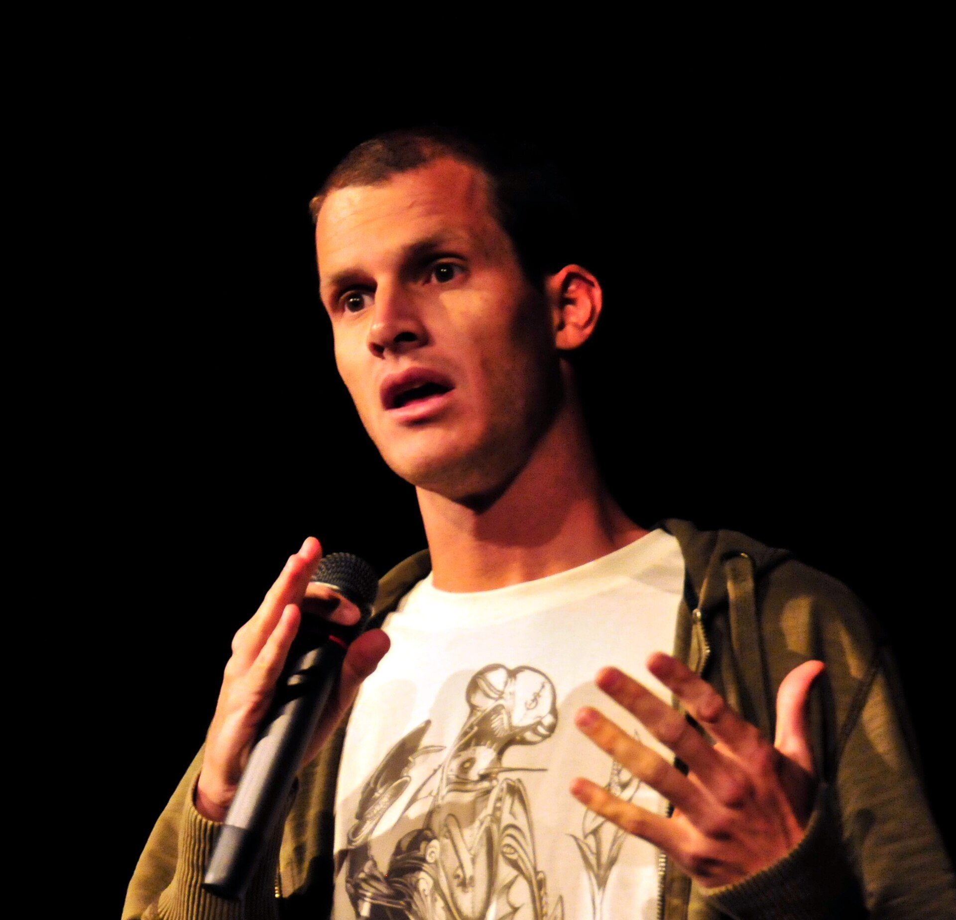 Daniel Tosh mentre fa stand-up su un palco buio nel 2008, illuminato dall'alto così che la parte superiore della testa e l'attaccatura dei capelli finiscono nell'abbaglio e nell'ombra.