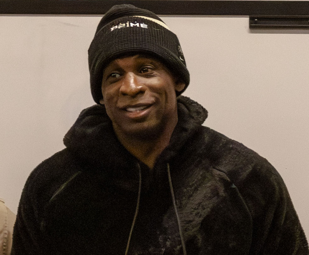 Deion Sanders in casa con un berretto nero e un cappotto con cappuccio, il berretto tirato giù in modo che non si vedano né capelli né attaccatura.