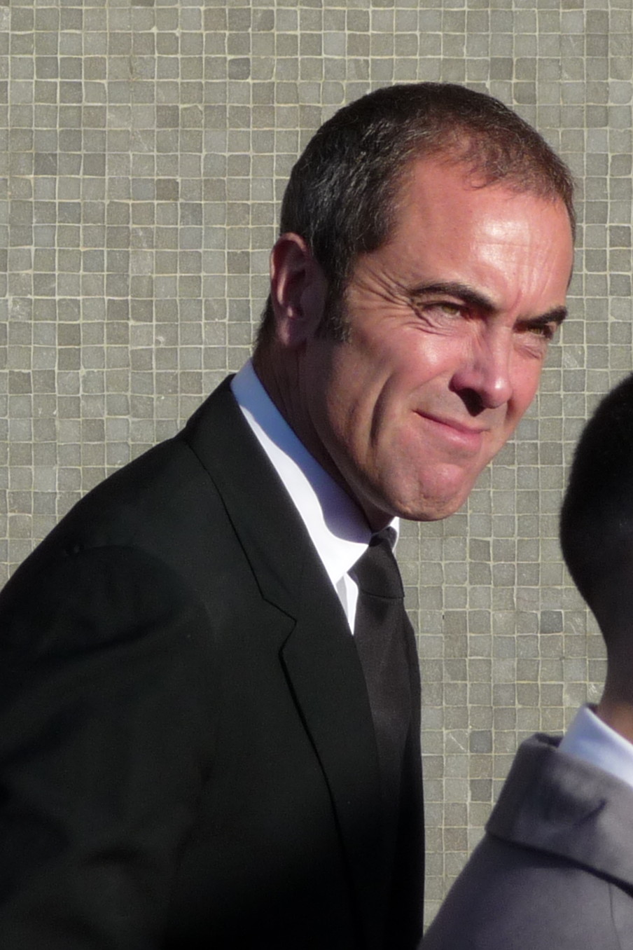 James Nesbitt all'aperto alla luce del giorno in un abito scuro, fotografato di tre quarti con i capelli corti e l'attaccatura frontale chiaramente illuminata.