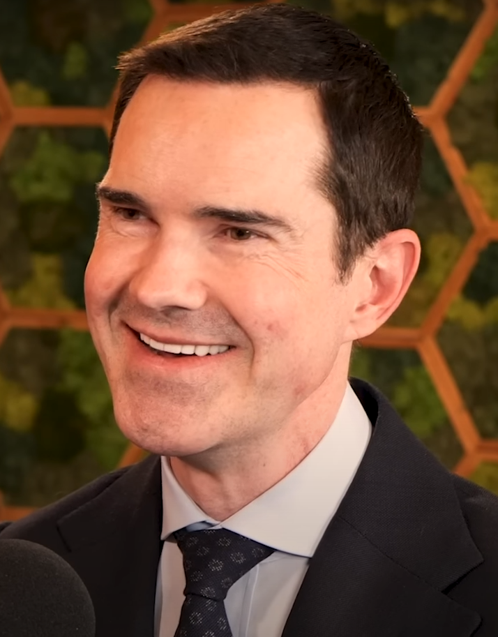 Jimmy Carr sorride in abito elegante a un evento all'aperto del 2024, i suoi capelli scuri pettinati all'indietro e l'attaccatura frontale visibile alla luce del giorno.