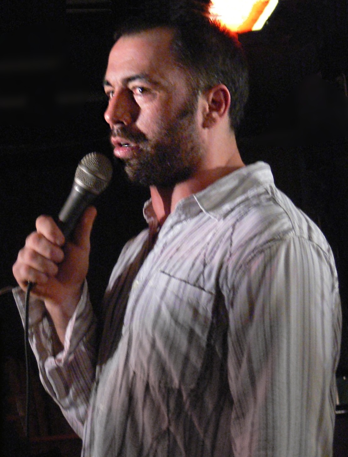Joe Rogan che fa stand-up su un palco di un club buio tenendo un microfono, illuminato da una singola luce dura dall'alto con i capelli tagliati molto corti.