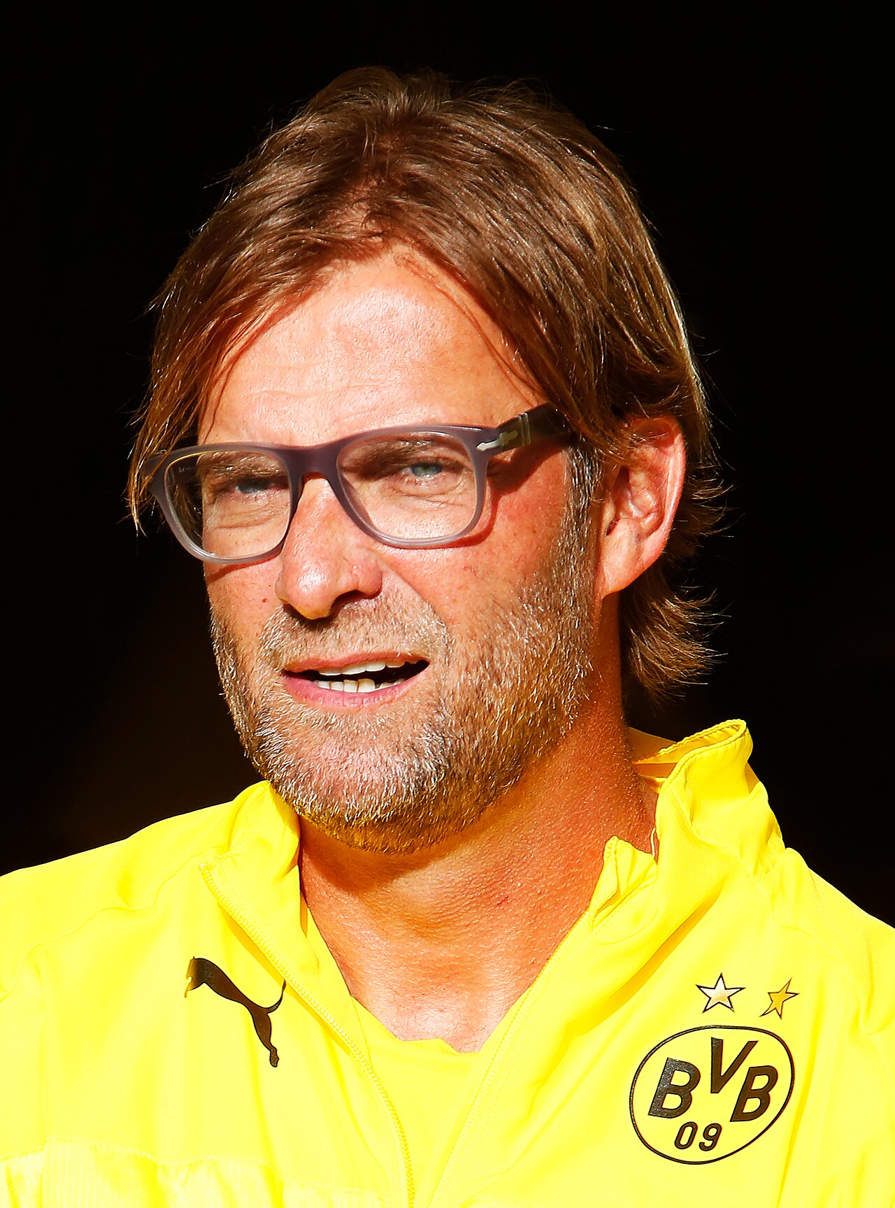 Jürgen Klopp in una giacca da allenamento gialla del Borussia Dortmund e occhiali nel 2014, con i capelli abbastanza lunghi da toccare il colletto.