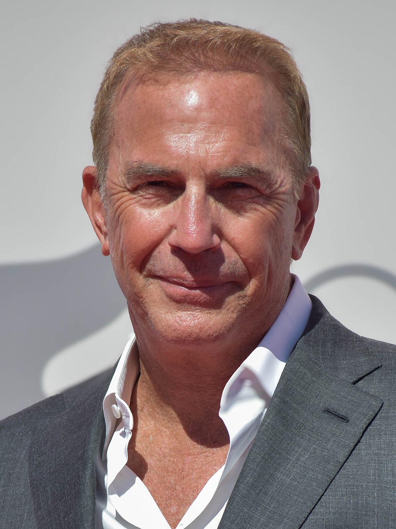 Kevin Costner a un festival cinematografico del 2024 con una giacca grigia, fotografato frontalmente con luce piatta da sala stampa e capelli spazzati sulla fronte.