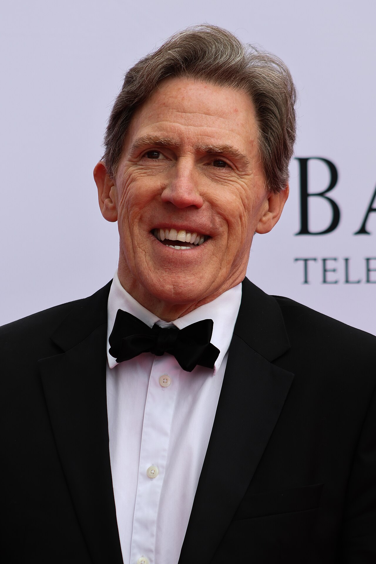Rob Brydon a un evento di premiazione nel 2026 in smoking e papillon, fotografato frontalmente con luce intensa dei flash, capelli grigi pettinati all'indietro e attaccatura frontale visibile.
