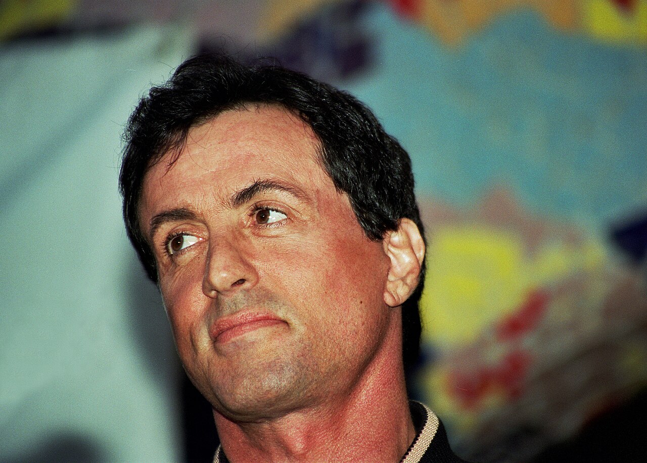 Sylvester Stallone photographed in three-quarter view at a press event, his dark hair worn thick and brushed back from the forehead.