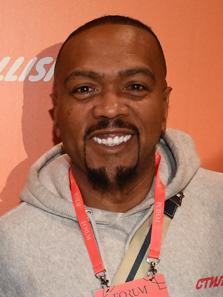 Timbaland con una felpa con cappuccio grigia e cordino da conferenza a un evento del 2019, fotografato da vicino e frontalmente su sfondo arancione con l'attaccatura frontale visibile.