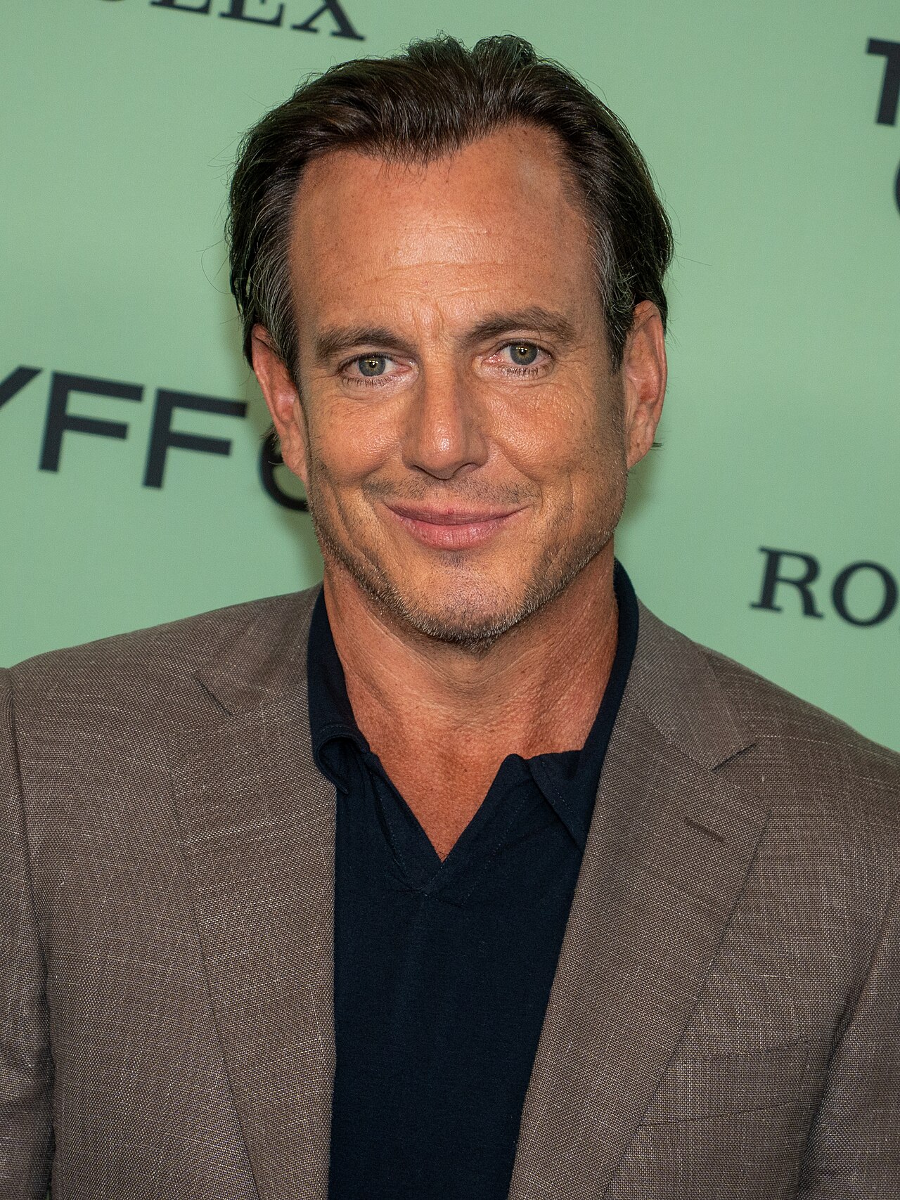Will Arnett at a 2025 film premiere in a tan jacket, photographed front-on under press lighting with his hair combed back off the forehead.